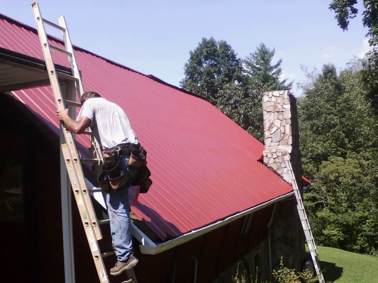 Roofer on a ladder beside a red metal roof and stone chimney — Soffit & Fascia Repair by Roofing Brothers in Palmerton